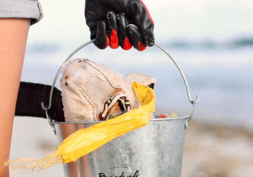 Person wearing red and black gloves holding a Boardwalk Boutique Hotel branded metal bucket filled with beach cleanup items on an Aruba beach