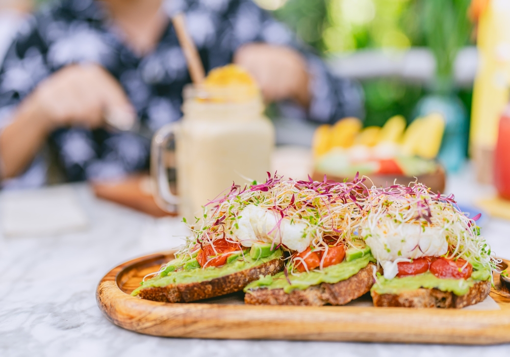 Avocado toast with poached eggs and microgreens served on a wooden board at Coco Café, with a smoothie in the background