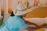 Hotel staff member in a blue uniform and sun hat arranging pillows on a bed, with a colorful bird mural in the background