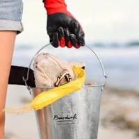 Person wearing red and black gloves holding a Boardwalk Boutique Hotel branded metal bucket filled with beach cleanup items on an Aruba beach