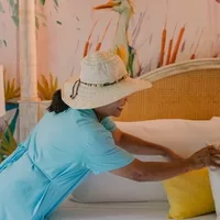 Hotel staff member in a blue uniform and sun hat arranging pillows on a bed, with a colorful bird mural in the background